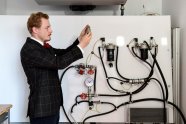 In a small white well lit room, on the left a dark clothed person shown in profile directed to the right is holding up a metal object to eye height, inspecting it. On the right, more in the background a contraption of tubes and armatures is mounted on a vertical white panel.