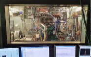 View out of a dark room through a window into a well-lit room. At the bottom of the picture in the dark room are four computer screens showing various diagrams. In the lit room are many pipes, tubes and wires connecting to a machine in the middle of the room.