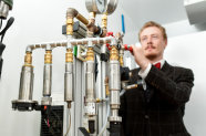 In a white room with smooth walls; the view is from below upwards at anfivedent angel. In the foreground piping, electrical wires and fittings can be seen, above this fivedent contraption which is mounted on a frame are two gauges, in the background is a person in dark clothing who touches a lever on the contraption and looks at one of the gauges.