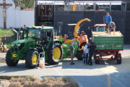 Im Vordergrund auf der linken Bildhälfte ein grüner Traktor mit davorstehendem Fass, mittig im Vordergrund ein Holzhäcksler welcher am Traktor angeschlossen ist, davor stehen drei Personen, auf der rechten Seite des Bildes im Vordergrund ein mit Hackschnitzeln gefüllter grüner Landwirtschaftsanhänger, auf dem sich zwei weitere Personen befinden, dahinter steht ein weiterer Traktor. Im Hintergrund befindet sich auf der linken Seite ein Grünstreifen welcher von einer Mauer abgeschlossen wird und auf der rechten Seite eine Halle mit Schiebetüren.