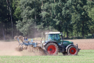 Fendt-Traktor grubbert von links nach rechts am Acker. Im Hintergrund ein Wald.