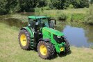 On a sunny day, in the foreground a green tractor with yellow wheels, which is parked on grassland near to a pond with some water plants on its surface. On the other shore of the pond are bushes and a wildflower meadow, in the background on the boundary of the meadow are bushes changing into woodland.