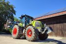 Hellgrüner Traktor mit roten Felgen steht nach rechts vor einer Scheune und hat einen Grubber und ein Frontgewichtangehängt.