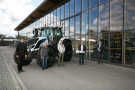 Fünf Männer stehen vor einem weißen Traktor mit scharzen Felgen, im Hintergrund ein Glasgebäude. Die zweite Person rechts überrecht der mittleren Personen eine überdimensionalen Schlüssel