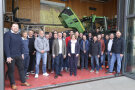 Etwa 20 Personen stehen in einer Halle mit seitlich geöffneten Glastüren vor einem grünen Traktor mit geöffneter Motorhaube.