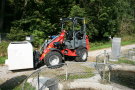 Ein roter elektrischer Hoftraktor transportiert einen weißen, ca. einen Kubikmeter großen Kanister auf Feldweg, im Vordergrund Fischbecken