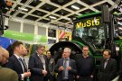 A in an exhibition hall a man talks to a surrounding audience. In the background is a John Deere tractor.