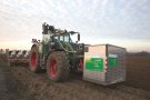 Fendt-Schlepper steht auf einem Acker mit einer großen Metallbox an vorderer Zapfwelle.