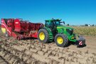 Ein John Deere-Traktor mit angehängtem Kartoffelroder fährt von links nach rechts am Kartoffelacker bei blauem Himmel.