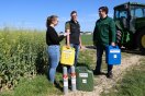 Eine Dame interviewt mit Klemmbrett zwei Herren. Sie stehen vor einem John Deere-Traktoren mit verschiedenfarbenen Kanistern und zwei kleinen Gasflaschen auf Feldweg.