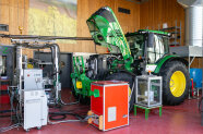 In the centre of a wood panelled hall with red flooring is a green tractor with yellow wheels, oriented slightly to the foreground, facing in left direction. The bonnet is opened upwards. The engine of the tractor is connected to a lot of wiring in different sizes. Multiple devices are surrounding the tractor to which the wires are leading. Behind the tractor is a blue cart, above which a silver exhaust duct is installed. In the back of it are shelves at the wall. On the back wall of the hall there is a poster of flowering rapeseed, some venting grilles and a double door.