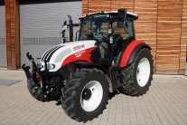 Ein rotweißer Steyr-Traktor steht nach vorne links schauend auf einem gepflasterten Hof vor einer hölzernen Betriebsgebäude.