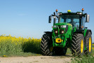 An einem sonnigen Tag, im rechten unteren Eck im Vordergrund unscharf ein Ackerrandstreifen, auf der rechten Bildhälfte ein John Deere 6135R Traktor neben einem auf der linken Bildhälfte befindlichen Rapsfeld in Vollblüte.