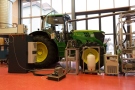 In a hall, in the foreground there are measurement devices and container, behind stands a green John Deere tractor which is connected to the measurement devices.