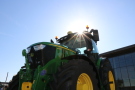 Im Vordergrund grüner John Deere 6250R Traktor von unten nach oben aufgenommen mit Sonne über dem Traktordach im Hintergrund.