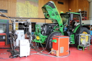 Picture of a tractor in a hall, in the foreground are various measuring devices, behind on the right connected to the measuring devices there is a green tractor with opened bonnet on the left are further measuring devices. In the background there can be seen shelfs and a poster with blooming rape.