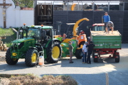 In the foreground of the left half of the picture there is a green tractor and in front of it a barrel in the middle is a woodchopper and three people in front of it, on the right is a farming trailer with to persons on top of it and another tractor behind. In the background on the left a grass strip with a wall behind it can be seen, on the right side a barn.