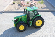 On a sunny day, picture taken at an angle downwards from high ground, shows the left side of a green tractor, which casts a shadow to the left lower corner of the picture, the tractor stands on cobble stones, in the background there is a rectangular area with gravel on which there are small trees and a table with benches.