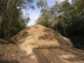 Das Bild zeigt Haufwerk von Waldhackschnitzeln bei Sonnenschein, welches konisch auf einem Lagerplatz im Wald aufgeschüttet ist. Die Lagerung findet unter freiem Himmel du ohne Abdeckung statt. Das Haufwerk ist über 5 m hoch. Im Hintergrund sind Bäume und ein blauer Himmel zu sehen.