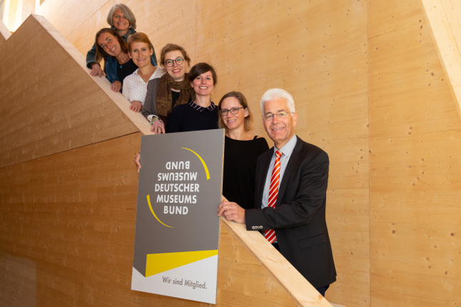 Team des NAWAREUM steht auf Holztreppe und präsentiert Schild mit Titel Deutscher Museumsbund