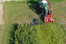 Eine große schwarze Drohne im Vordergrund fliegt über eine Silphieparzelle. Vor der Parzelle steht ein grüner Traktor mit angebautem roten Häcksler zur anschließenden Ernte.