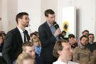 Zwei Männer, einer mit einem Mikrofon in der Hand, stehen zwischen der Zuhörerschaft und stellen Fragen
