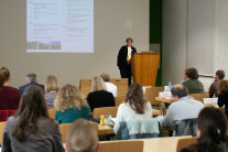 Eine Rednerin steht an einem Pult und spricht zu Publikum