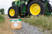 Ein 5 Liter Kanister gefüllt mit gelber Flüssigkeit steht auf einem Feldweg vor einem unscharf fotografierten John Deere-Traktor. links und rechts Gräser